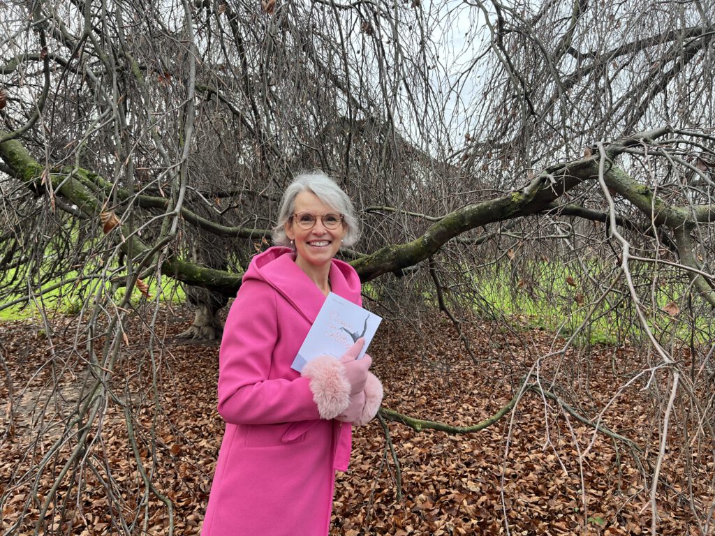 Katrien Jacobi steht im pinkfarbenem Mantel mit ihrem Buch lächelnd vor einem winterlich blattlosen Baum