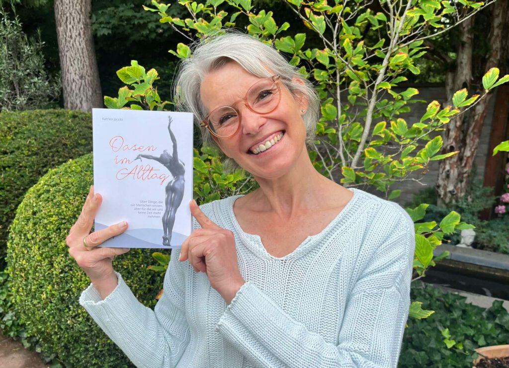 Dr. Katrien Jacobi, graue Haare, Brille, hält lächelnd ihr Buch Oasen im Alltag in die Kamera